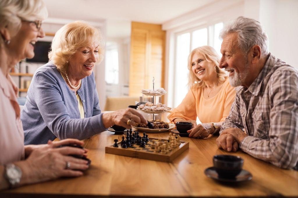 Four older friends laughing together over coffee and a game of chess at a kitchen table