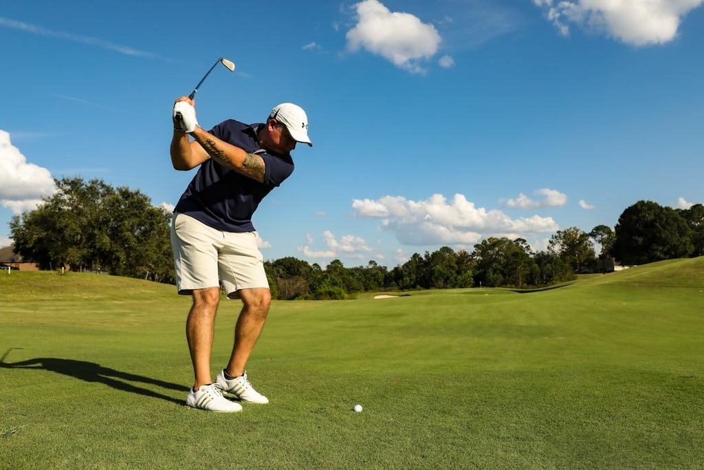 A man in a dark polo shirt and white shorts swinging a golf club on a sunny course