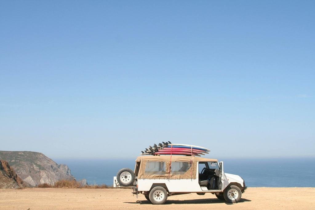 A white off-road vehicle with surfboards on its roof, parked on a clifftop above the sea