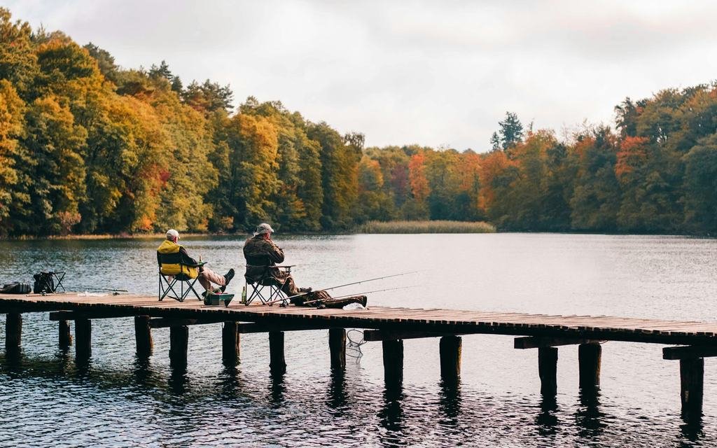 Two people sitting in folding chairs on a wooden jetty, fishing on a lake lined with autumn trees