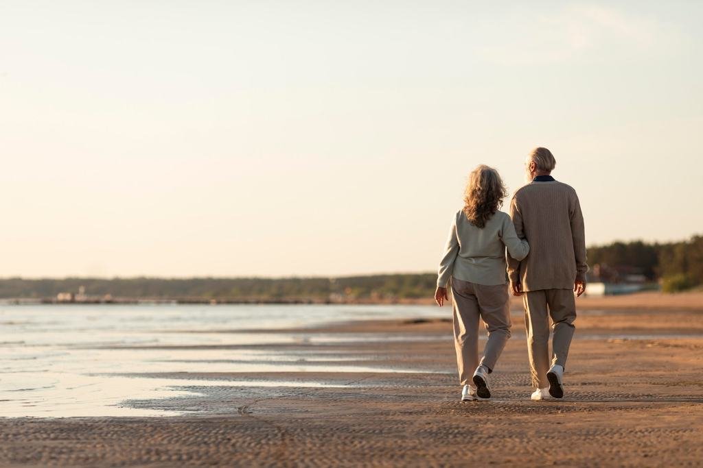 An older couple walking arm in arm along a sandy beach in soft evening light